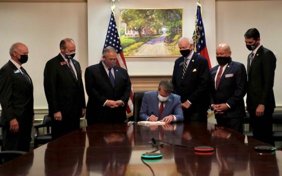 Men standing around a table where governor is seated signing a bill, under a painting of a slave plantation