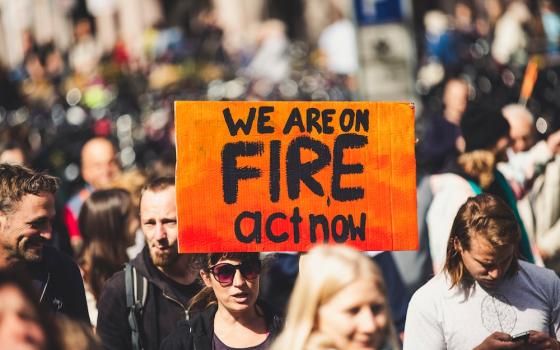 Demonstrators marching together, while a person holds a sign that reads "We are on fire, act now." (Unsplash/Markus Spiske)