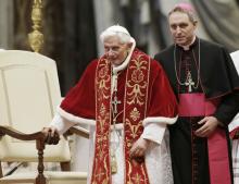 A white bishop stands behind Pope Benedict XVI who is wearing red and gold vestments