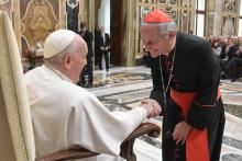 A cardinal leans down to greet a seated Pope Francis in a Vatican reception hall