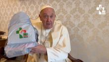 Pope Francis holds up a gray backpack with the World Youth Day logo as he speaks in a room with the Vatican City emblem on the cream wallpaper