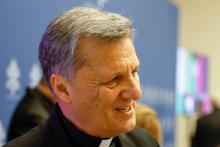 A gray-haired white man wearing a clerical collar smiles. The Vatican press conference backdrop is blurry in the background.