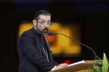 A light-skinned man with a beard and glasses and black hair speaks into a microphone at a podium