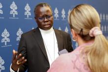 A Black man wearing glasses and a white collar talks to a blond woman who holds a phone towards him