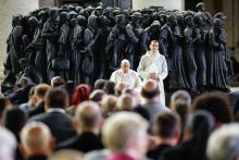 Pope Francis and a priest wearing white stand in front of metal statue of a mass of people. A crowd of people stands on the other side of them.