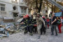 Decimated street, covered in rubble; uniformed emergency workers carry stretcher through wreckage.