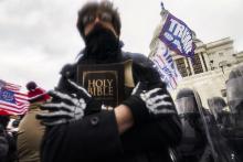 Rioter with face obscured, holds bible to chest, in the background is Capitol building dome and various flags, including one bearing former president Donald Trump's last name.