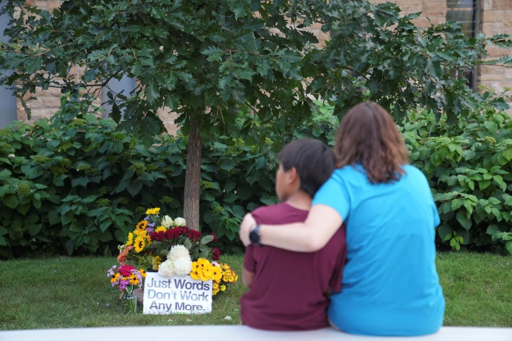 People sit across from a memorial near boarded windows damaged in the Aug. 27 fatal school shooting at Annunciation Catholic Church.