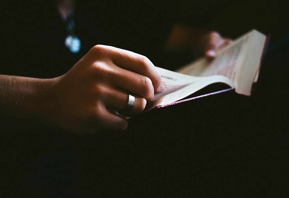 A dark photo illustration features a dramatically lit close-up of hands turning the page of a Bible. (Unsplash/Anna Hecker)