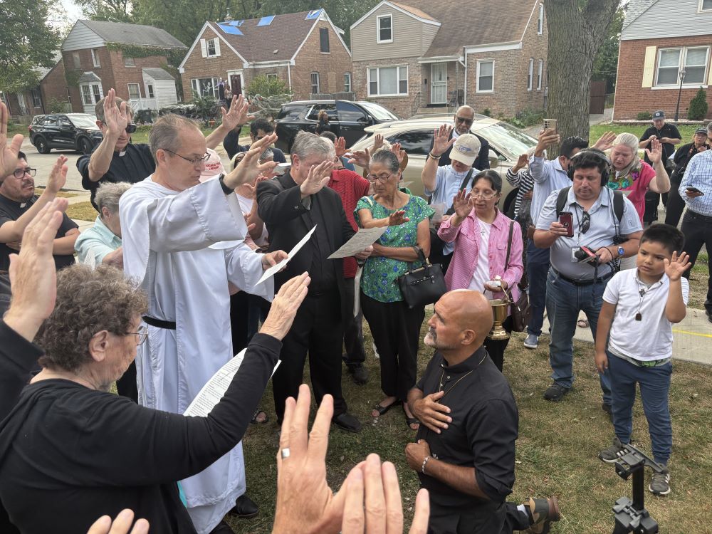 Fr. Gary Graf kneels outside while people raise their hands to bless him. pporters and friends outside Pope Leo XIV's childhood home in Dolton, Illinois, on the first day of his two-month walk, Oct. 6. (Courtesy of Lauren Foley)