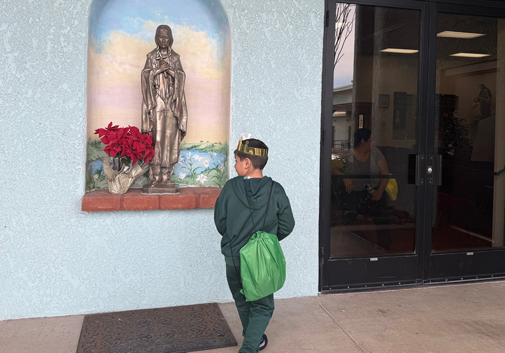 A young migrant boy wearing a cardboard crown walks by a statue of St. Kateri Tekakwitha, the first Native American woman saint who lends her name to a parish in Tucson, Arizona, where a Three Kings Day celebration was organized Jan. 4, 2026. (Anita Snow)