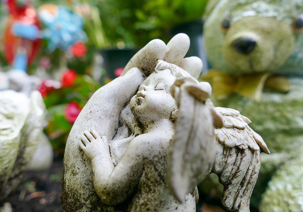 A photo illustration shows a sculpture of an angel child resting in adult hands, pictured in an outdoor setting, perhaps a cemetery. (Pixabay/dominic_winkel)