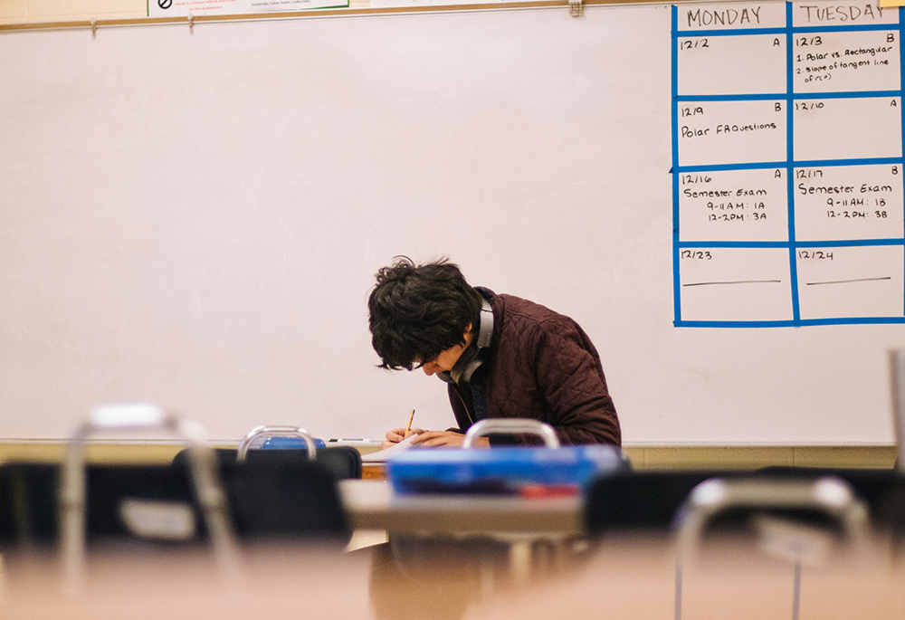 A young man works in a classroom setting. He is seated amid tables and chairs, with a dry erase board behind him. (Unsplash/Jeswin Thomas)