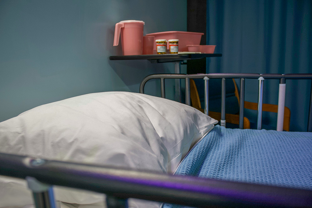 An empty hospital bed with a white pillow and a blue blanket, with an empty chair next to it. (Unsplash/Bret Kavanaugh)