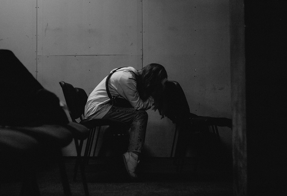 A lone person seats with their head in their arms, in an empty row of seats pictured in a black-and-white photo illustration. (Unsplash/Tymofii Tarasov)