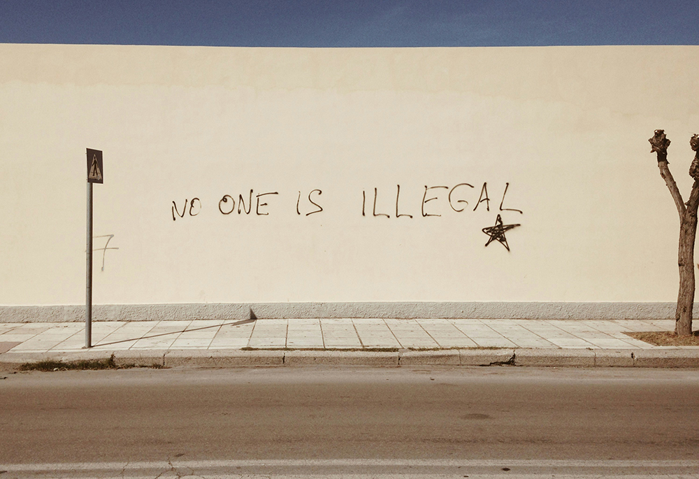 The message "No one is illegal" is spray-painted on a tall wall that runs the length of a sidewalk in an outdoor setting. (Unsplash/Miko Guziuk)