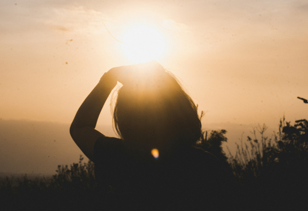 A silhouette of a woman who holds her hand up as she looks to the sunset in an outdoor setting with plants and a wide horizon. (Unsplash/arief santoso)