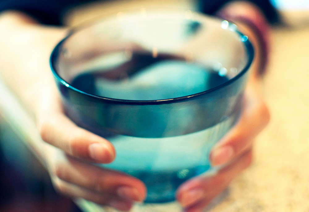A photo presents an extreme close-up of a pair of hands holding and possibly offering forward a full glass of drinking water. (Unsplash/Amanda Yum)