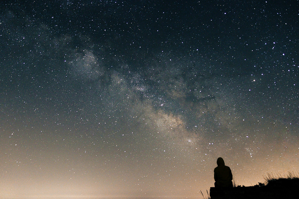 Silhouette of a person sitting on a hill looking at the Milky Way galaxy (Unsplash/Clement Duguerre)