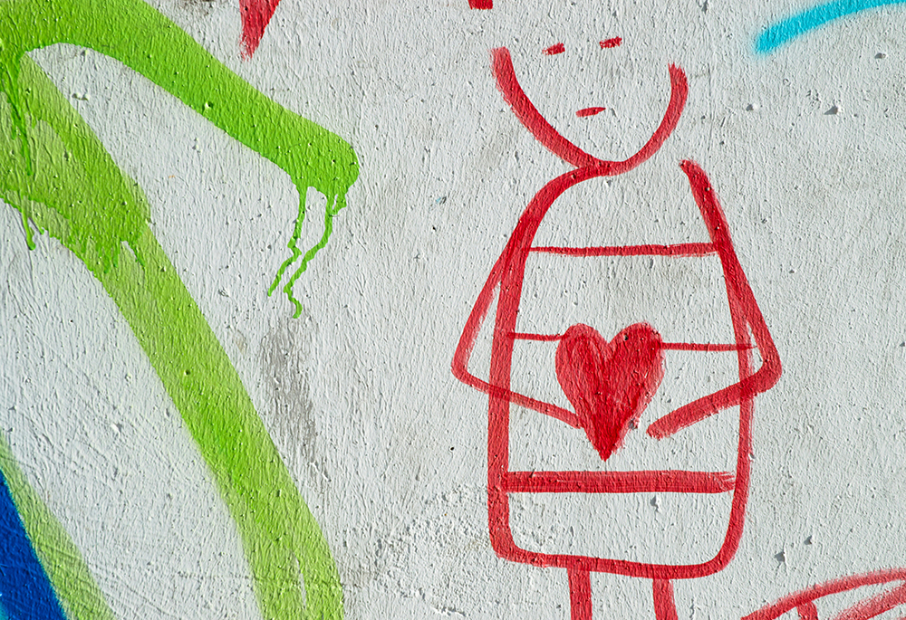 A photo illustration shows a close-up shot of a line drawing made on what looks to be a blank, white wall. A person, drawn in red, wears a blank expression as they hold a heart, which is also drawn and colored in red, in a traditional, symmetric heart shape. A solid line of green and some blue appear to the figure's left on the wall. (Unsplash/Eduardo Goody)