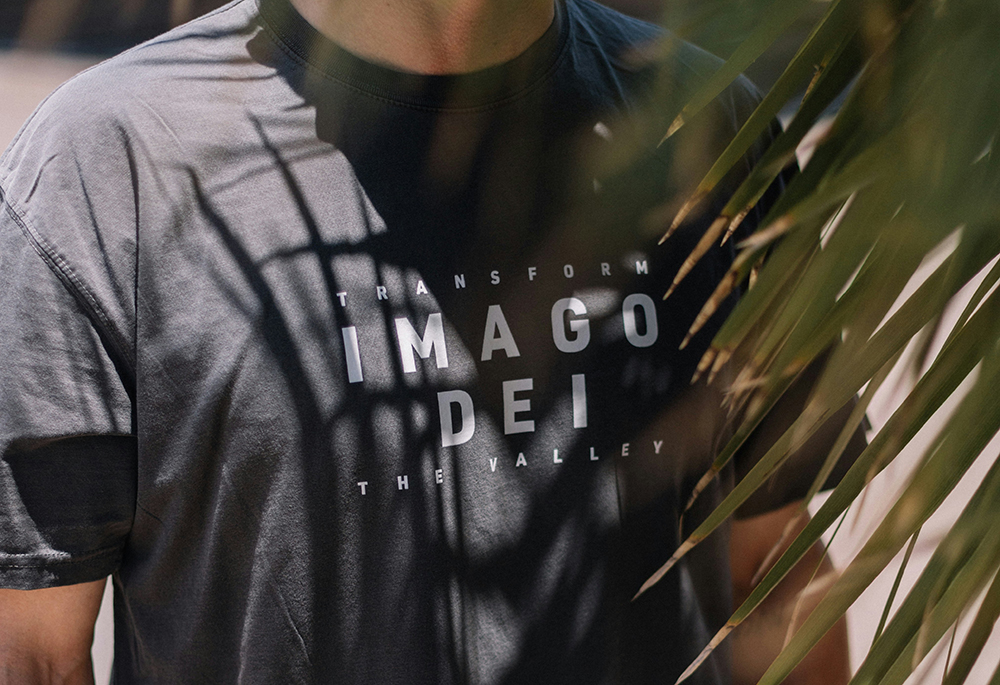 A man wears a T-shirt that displays the words "imago Dei" and "transform the valley" in all caps, white text against black cloth. He stands next to a palm tree. (Unsplash/Phillip Goldsberry)