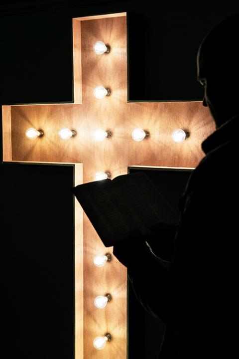 A person reads a book in front of an electronic cross lit by bulbs in a darkened room. (Unsplash/Mark Stuckey)