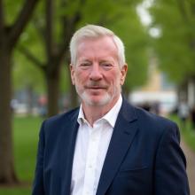 A photo of John Berry in a white collared shirt and navy blue jacket with out-of-focus trees in the background