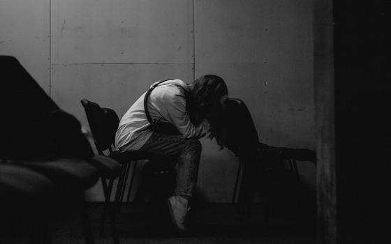 A lone person seats with their head in their arms, in an empty row of seats pictured in a black-and-white photo illustration. (Unsplash/Tymofii Tarasov)