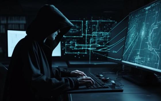 A hooded figure sits in dark room in front of lit-up computer