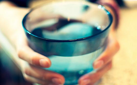 A photo presents an extreme close-up of a pair of hands holding and possibly offering forward a full glass of drinking water. (Unsplash/Amanda Yum)