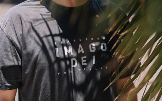 A man wears a T-shirt that displays the words "imago Dei" and "transform the valley" in all caps, white text against black cloth. He stands next to a palm tree. (Unsplash/Phillip Goldsberry)